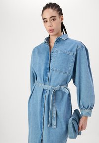 Denim srajčna obleka v svetlo modri barvi, ima ovratnik, dolge napihnjene rokave ter pasni trak. Žep na prsih in gladka, teksturirana tkanina.