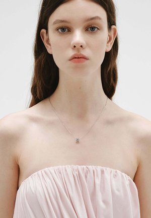 Joven con cabello largo y castaño que lleva un vestido sin tirantes de color rosa pálido y un delicado collar de plata con un pequeño colgante.