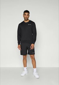 Un homme se tient contre un fond neutre, portant un sweat à capuche noir Champion, un short noir, des chaussettes blanches mi-mollet et des baskets blanches.