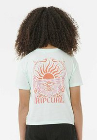 Persona con cabello rizado de espaldas, wearing una camiseta azul clara con un gráfico de un sol y una ola en rosa y naranja, y el texto "RIP CURL".