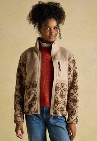 Woman with curly hair wears a beige patterned fleece jacket over a red knit sweater and blue jeans, standing against a neutral background.