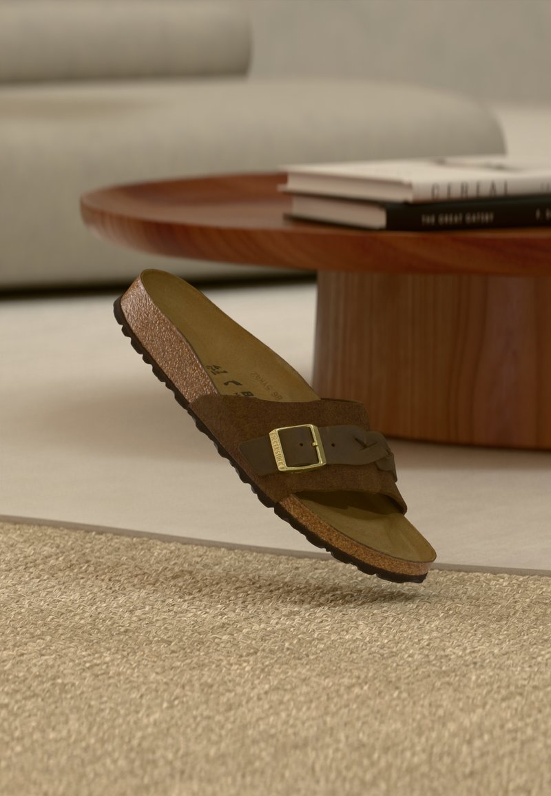 Καφέ σουέντ σανδάλι με ρυθμιζόμενο λουράκι και χρυσή αγκράφα, τοποθετημένο πάνω σε μπεζ χαλί και φόντο από ξύλινο τραπεζάκι σαλονιού.