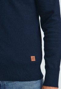 Sötétkék, hosszú ujjas pulóver bordás mandzsettákkal és aljjal, világoskék farmernadrág mellett viselve, amelyen egy kis barna bőrból készült márkajelzés található az alján.