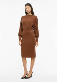 Brązowa dzianinowa sukienka z dekoltem typu łódka, długimi balonowymi rękawami i dopasowaną talią, zestawiona z czarnymi butami na wysokim obcasie z złotymi akcentami.