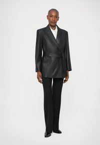 Čierny kožený oversize blejzer s opaskom v páse, ostrými klopami a dlhými rukávmi, skombinovaný s upravenými čiernymi nohavicami a špicatými topánkami.