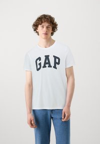 Balta medvilninė marškinėliai su dideliu juodu "GAP" logotipu priekyje. Jie turi klasikį apvalų kaklą ir trumpas rankoves.