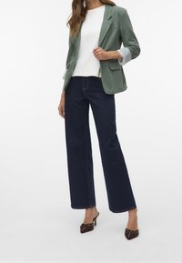 Femme portant un blazer vert avec des manches à rayures retroussées, un top blanc, un jean noir taille haute à jambes larges et des mules à talons hauts marron.