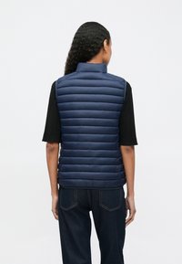 Osoba s tamnom kovrčavom kosom koja nosi tamnoplavu podstavljenu prsluk, crni majicu kratkih rukava i tamnoplave traperice, prikazana s leđa.