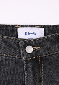 Μαύρο τζιν παντελόνι από denim με ετικέτα Rivvia, που διαθέτει ασημένιο κουμπί και χρυστή ραφή κατά μήκος των ραφών. Κοντινή λήψη στην περιοχή της μέσης.