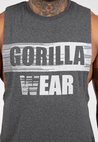 Grijze mouwloze tanktop van katoen; heeft vet zwart-witte tekst: "GORILLA" boven "WEAR" met gestreepte accenten op de letters.