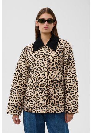 Vrouw draagt een jas met luipaardprint met een grote zwarte kraag, donkere zonnebril en blauwe jeans, staand tegen een effen lichte achtergrond.