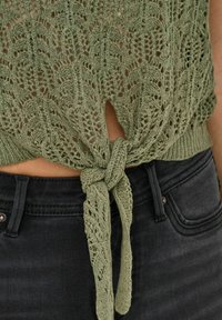 Top in maglia verde oliva con motivo di pizzo aperto e dettaglio di fiocco in vita, abbinato a jeans neri con tasca anteriore e chiusura con bottone.