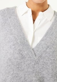 Grauer Strickwesten mit tiefem V-Ausschnitt, über einem weißen Hemd mit Kragen getragen. Die Textur ist weich und leicht flauschig.