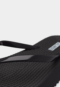 Sandale tongs noire avec semelle texturée et fines lanières brillantes, vue en gros plan sur la partie avant supérieure.