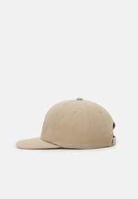 Boné de beje com uma coroa arredondada, aba curva, ilhos para ventilação e uma pequena etiqueta de marca de lado.