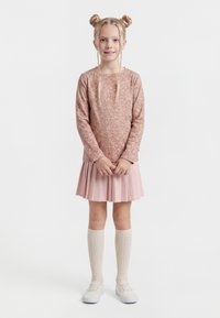 Mladá dívka se světlými vlasy upravenými do dvou drdolů a copy, oblečena v růžovém květovaném tričku s dlouhými rukávy, růžové plisované sukni, krémových podkolenkách po kolena a bílých botách.