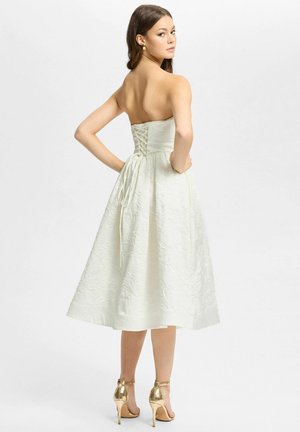 Vrouw in een witte, strapless midi-jurk met textuur en veterdetail aan de achterkant, poseert met handen in de zij, draagt gouden hoge hak sandalen.