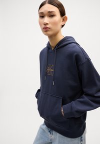 Sweatshirt azul marinho com capuz em tecido de algodão, com bolso frontal, apresentando um logo laranja "Calvin Klein" e cordões ajustáveis.