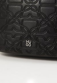 Geantă din piele texturată neagră, cu un model embosat de bucle și un accent cu logo-ul argintiu "HUGO". Finisaj neted cu un design modern.