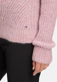 Hellrosa gestrickter Pullover mit lockerer Passform, gerippten Bündchen und einem silbernen Akzentdetail. Weiche Textur mit leicht flauschigem Aussehen.