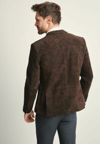 Brązowa zamszowa marynarka o dopasowanym kroju, z wycięciem na klapach i rozcięciem z tyłu, stylizowana z białą koszulą i ciemnymi spodniami.