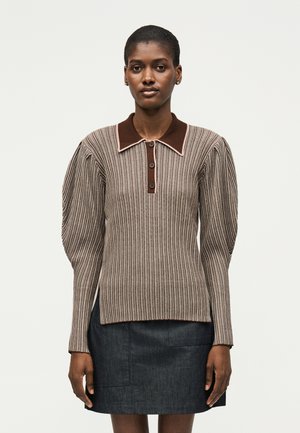 Mladá žena nosi ribovaný pásikavý sveter s naberanými rukávmi, hnedým golierom a gombíkmi, skombinovaný s tmavou rifľovou sukňou.