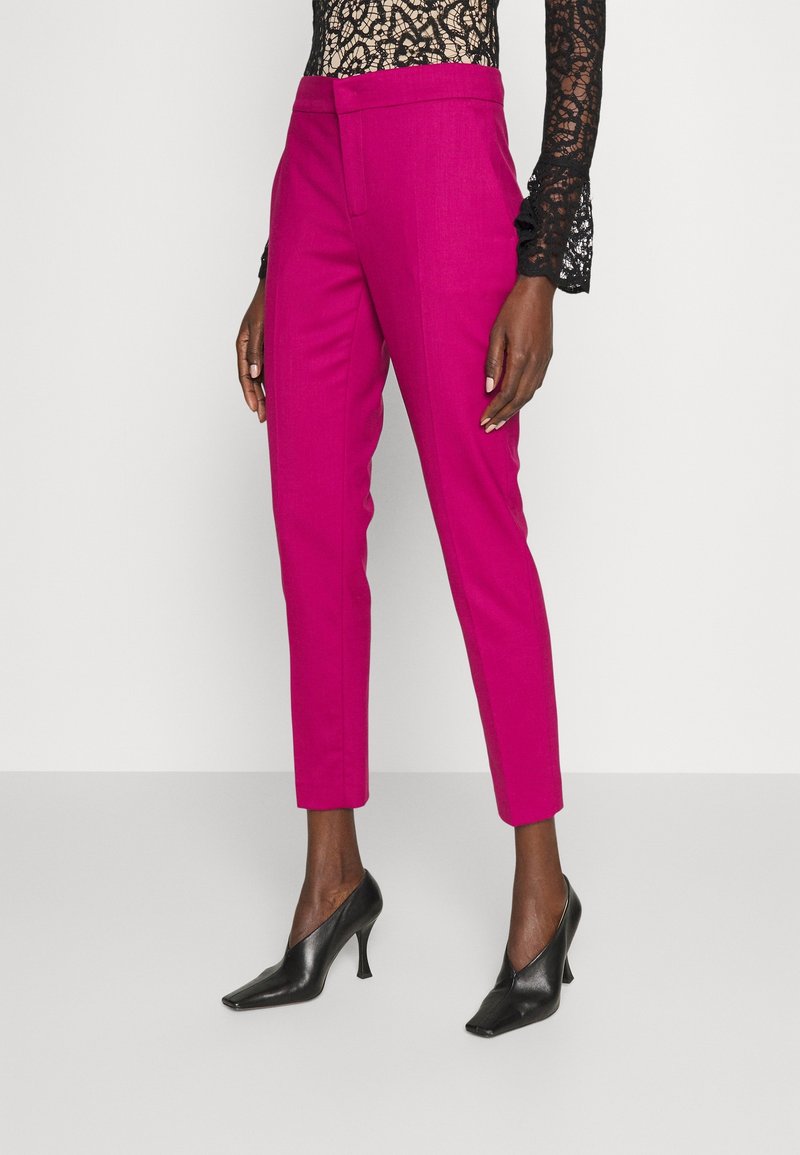 Pantalon fuselé rose vif jusqu'à la cheville porté avec des escarpins noirs à bout pointu et un haut en dentelle noire à manches longues.