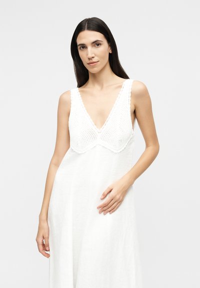 Nő hosszú sötét hajjal, fehér, ujjatlan ruhában, amelynek kötött V-nyakú felsőrésze van, egyszínű világos háttér előtt.