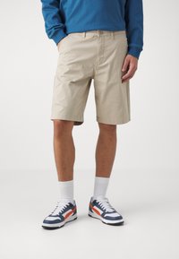 Beige Shorts mit flacher Vorderseite, seitlichen Taschen und Knopfverschluss. Getragen mit einem blauen Langarmshirt und blauen, weißen und orangefarbenen Sneakers.