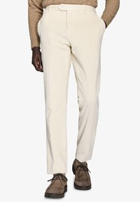 Pantaloni in velluto a coste color crema con una finitura testurizzata, caratterizzati da tasche laterali e una vestibilità slim, abbinati a calzature marroni.