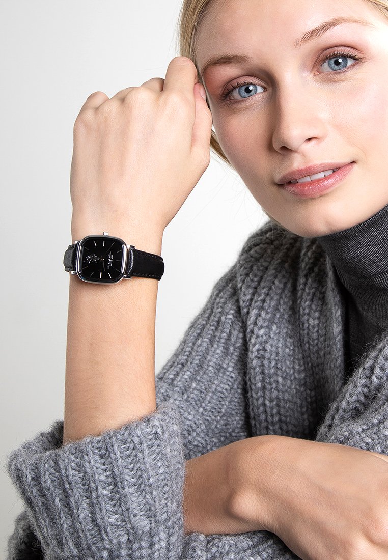 Quadratische schwarze Uhr mit silbernen Akzenten, an einem schwarzen Lederarmband befestigt, am Handgelenk getragen, neben einem grauen Strickoberteil.