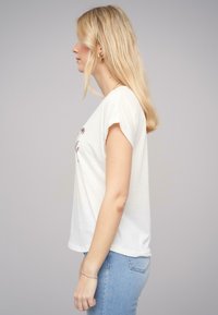 T-shirt blanc à manches courtes avec un imprimé floral, en coton doux. Porté avec un jean bleu clair, mettant en valeur une coupe décontractée et un tombé léger.