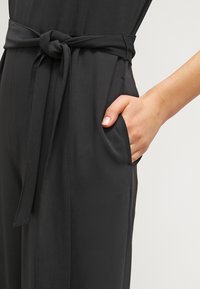 Svart jumpsuit med knytmidja, sidfickor och en mjuk textur. Tyget verkar lätt och faller elegant.