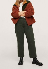 Fleecejakke i orange og grøn med høj krave, sammen med olivengrønne bukser og sorte kraftige støvler. Teksturerede stofdetaljer synlige.