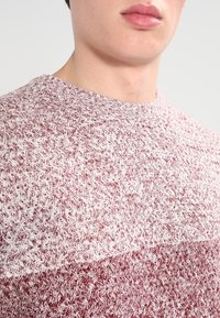 Teksturoitu neulepaita, jossa on punavalkoinen heather-kuvio, ribattua pyöreää kaula-aukkoa ja lyhyet hihat. Lähikuvassa korostuvat kuviot.