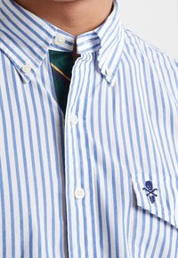 Skjorta med krage i ljusblå och vita vertikala ränder, med ett litet broderat skalle och korslagda ben på vänster bröstficka.