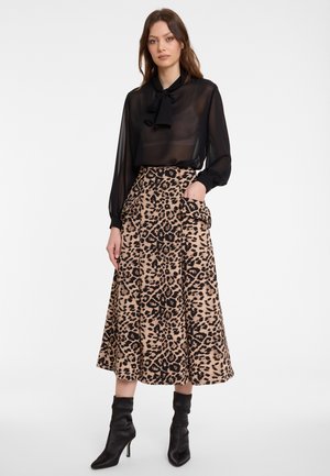 Frau in einer durchsichtigen schwarzen Bluse mit langen Ärmeln und Schleifenkragen, einem midi Rock mit Leopardenmuster und Klappentaschen sowie schwarzen, hohen Stiefeletten, die steht.