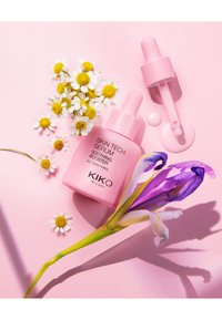 Botella rosa de suero KIKO Skin Tech con cuentagotas, acompañada de margaritas blancas y un iris morado, sobre un fondo rosa claro.