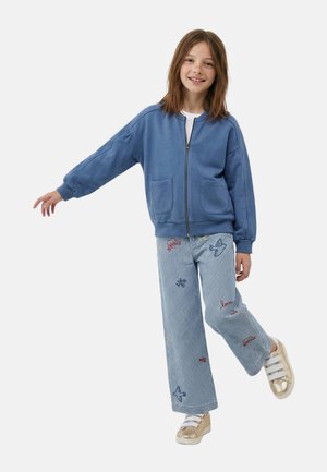 Fille souriante en équilibre sur une jambe, portant une veste bleue à fermeture éclair, un pantalon rayé avec une broderie d'avion et des baskets dorées.