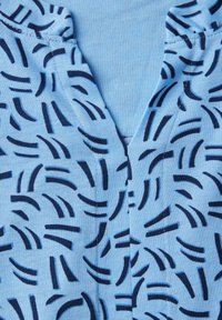 Lichtblauw top gemaakt van zachte stof met een patroon van donkerblauwe gebogen lijnen. Het ontwerp heeft een V-halslijn en subtiele stikseldetails.