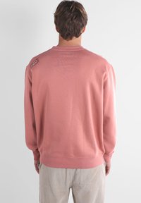 Sweat-shirt rose en tissu doux, avec un col rond et des poignets côtelés. Comprend un petit logo sur l'épaule gauche.