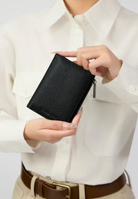Portefeuille en cuir noir texturé tenu dans une main, avec une fermeture éclair et une petite étiquette en métal, sur un fond de chemise clair.