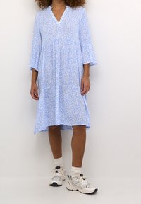 Světle modré květinové šaty s výstřihem do V, tříčtvrtečními zvonovými rukávy a siluetou do A, doplněné bílými a šedými teniskami.