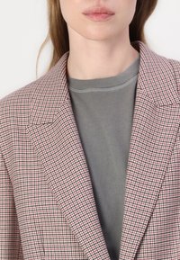 Rosa og svart houndstooth-blazer med skarpe revers, over en grå genser. Stoffet er teksturert med en skreddersydd passform og rene linjer.