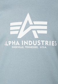 Ljusblå bomulls-t-shirt med en framträdande vit logotyp med tre horisontella ränder och texten: "ALPHA INDUSTRIES, KNOXVILLE, TENNESSEE, U.S.A."