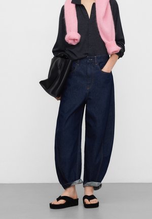 Losse denim jeans in donkerblauw met opgerolde boorden, gecombineerd met een zwarte handtas en een roze trui om de nek gedrapeerd. Zwarte sandalen.