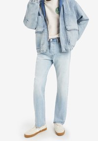 Jeans de mezclilla azul claro con un corte recto, combinados con una chaqueta azul claro y una sudadera crema con un gráfico verde. Zapatillas blancas.