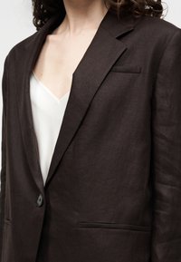 Ženska, oblečena v temno rjav blazer z vrezanimi ovratniki in enim črnim gumbom, čez bel top z V-izrezom.