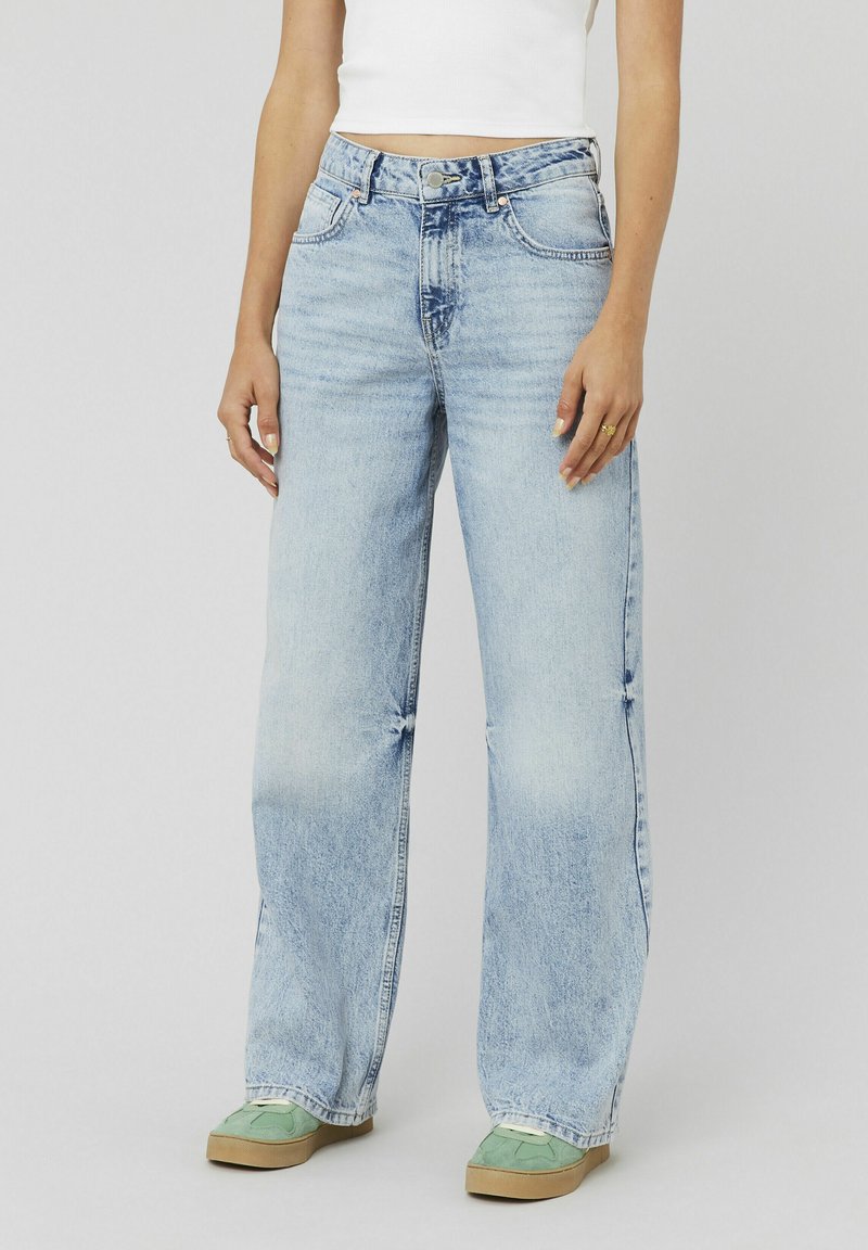 Femme portant un jean large bleu clair, un débardeur blanc court et des baskets vertes sur un fond gris uni.