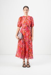 Woman wearing a red floral midi dress with puff sleeves, holding a light purple handbag, and wearing black strappy sandals.